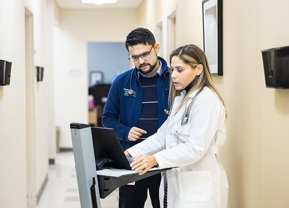 A male and female healthcare working looking at a laptop on a rolling stand in a hallway