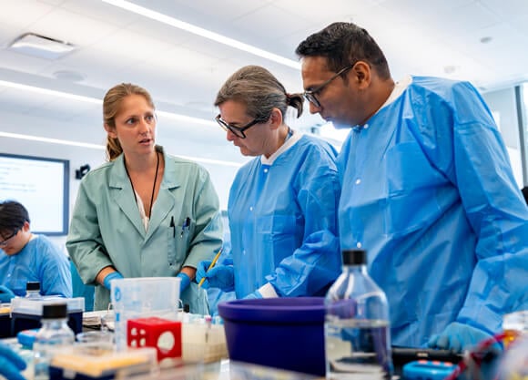 A faculty member and two adult students in medical wear at a lab counter.