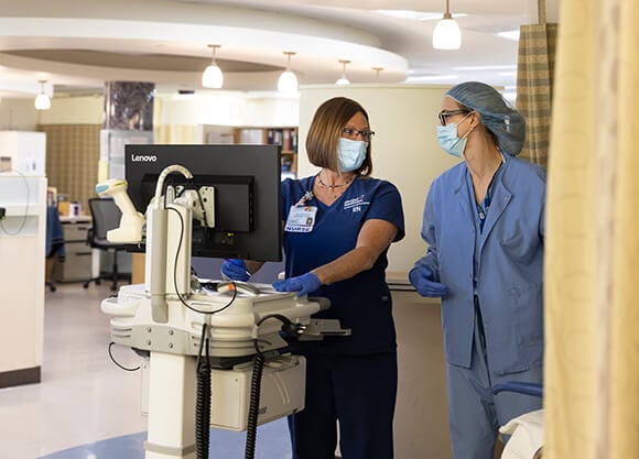 A nurse in front of a mobile laptop station talking to a doctor in surgical gear