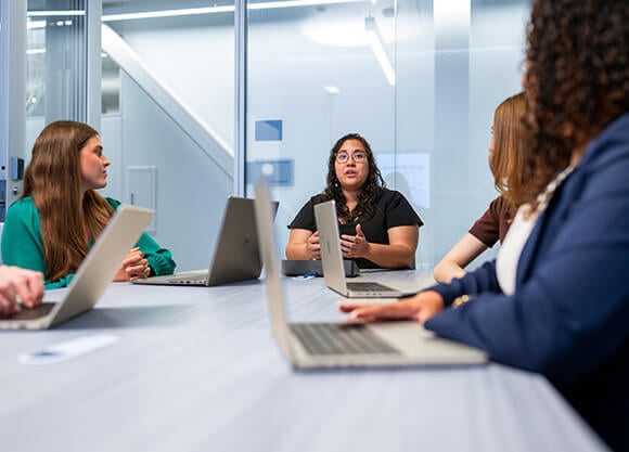 A woman at the head of a table talking during a meeting with 3 other women out of focus