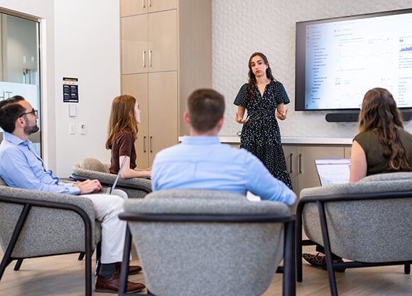 Participants seated in a meeting space listen to a presenter standing in front of a wall-mounted screen displaying charts and performance data during a group discussion.