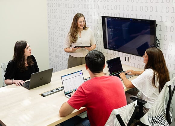Participants work around a conference table using laptops while one participant stands at the front of the room presenting from a tablet beside a wall-mounted display showing computer code.